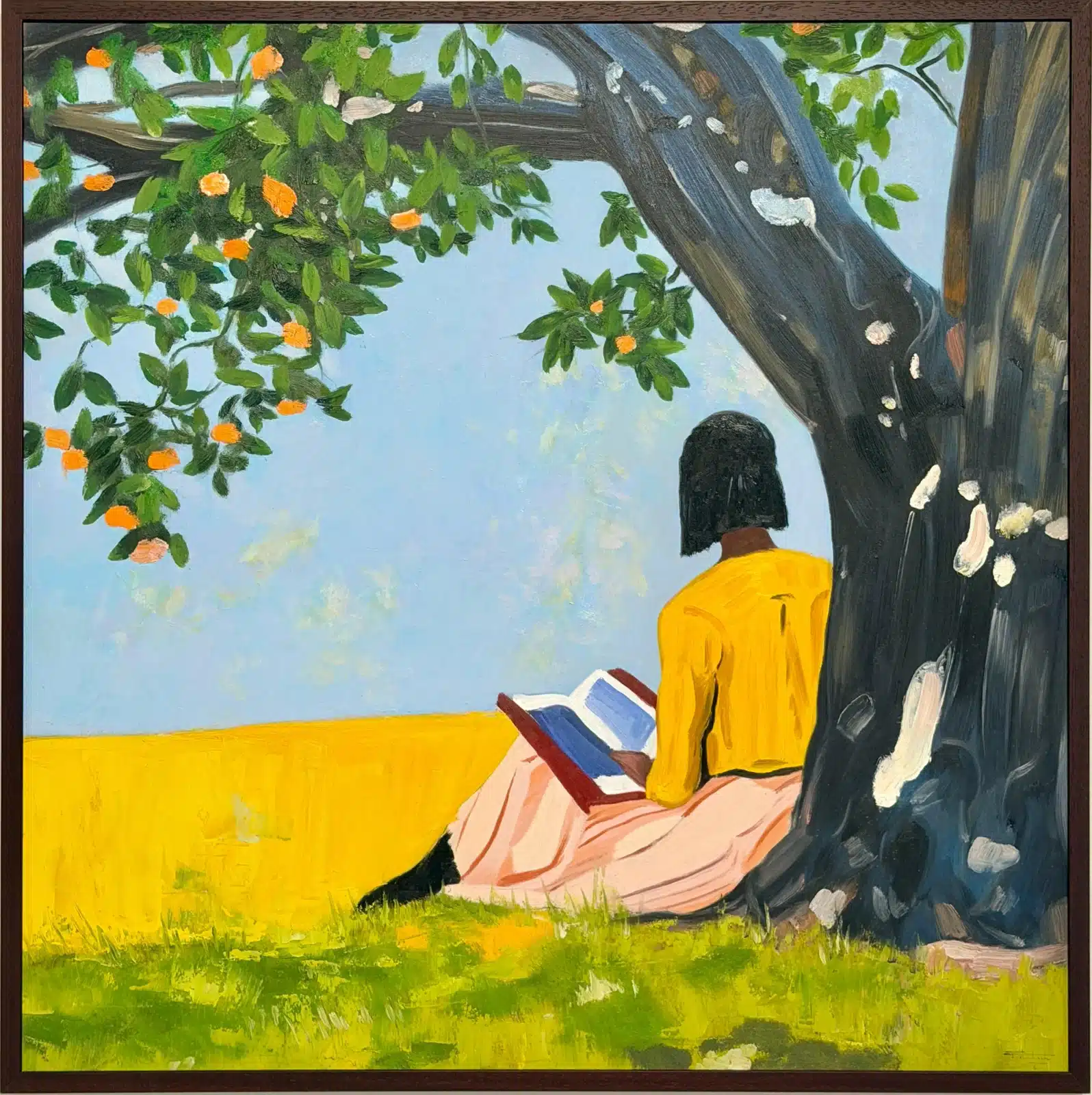 Une femme en pull jaune lit, adossée à un grand arbre au bord d’un champ doré.